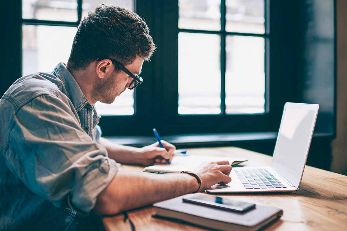Person sits at a table with a laptop writing in a notebook.